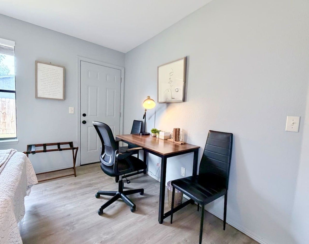 A small, neatly arranged room with a desk, two chairs, a lamp, and a bed.