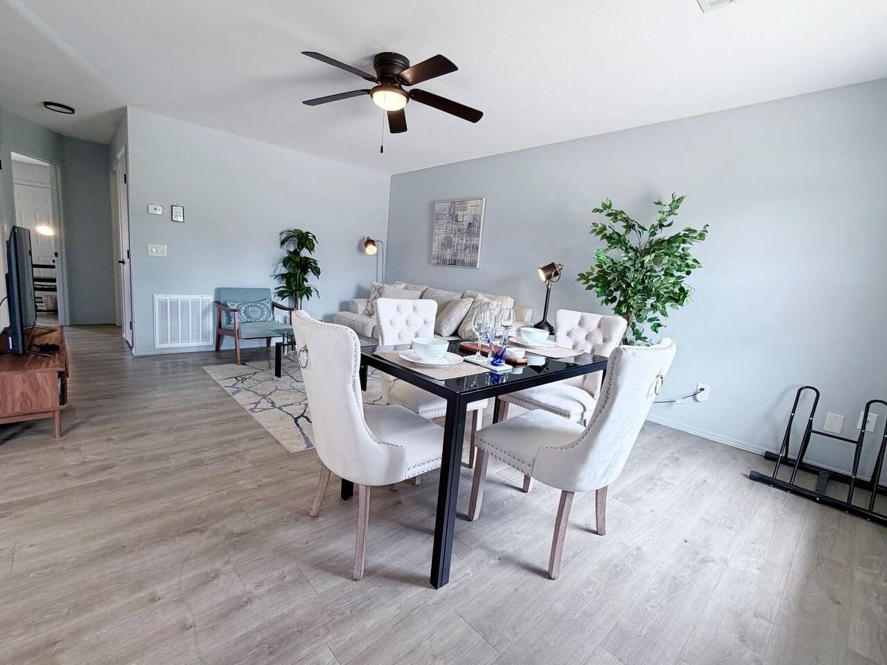 A modern dining area with a glass-top table, white upholstered chairs, and a ceiling fan.