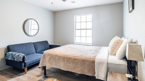 A bedroom with a bed, a blue sofa, a round mirror, a ceiling fan, and a window.