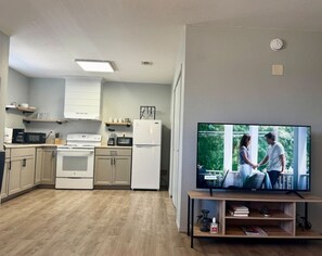 A modern kitchen with white appliances, a dining area with a TV, and a view of greenery outside.