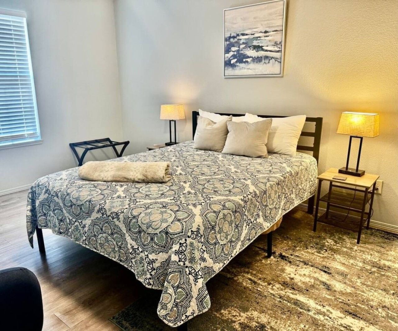 A bedroom with a patterned bedspread, two bedside lamps, a headboard, and a framed picture on the wall.
