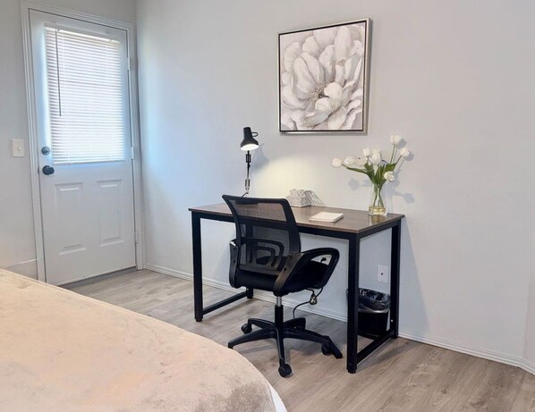 Une chambre à coucher avec un lit, un bureau avec une chaise, une lampe, un tableau encadré et un vase avec des fleurs.