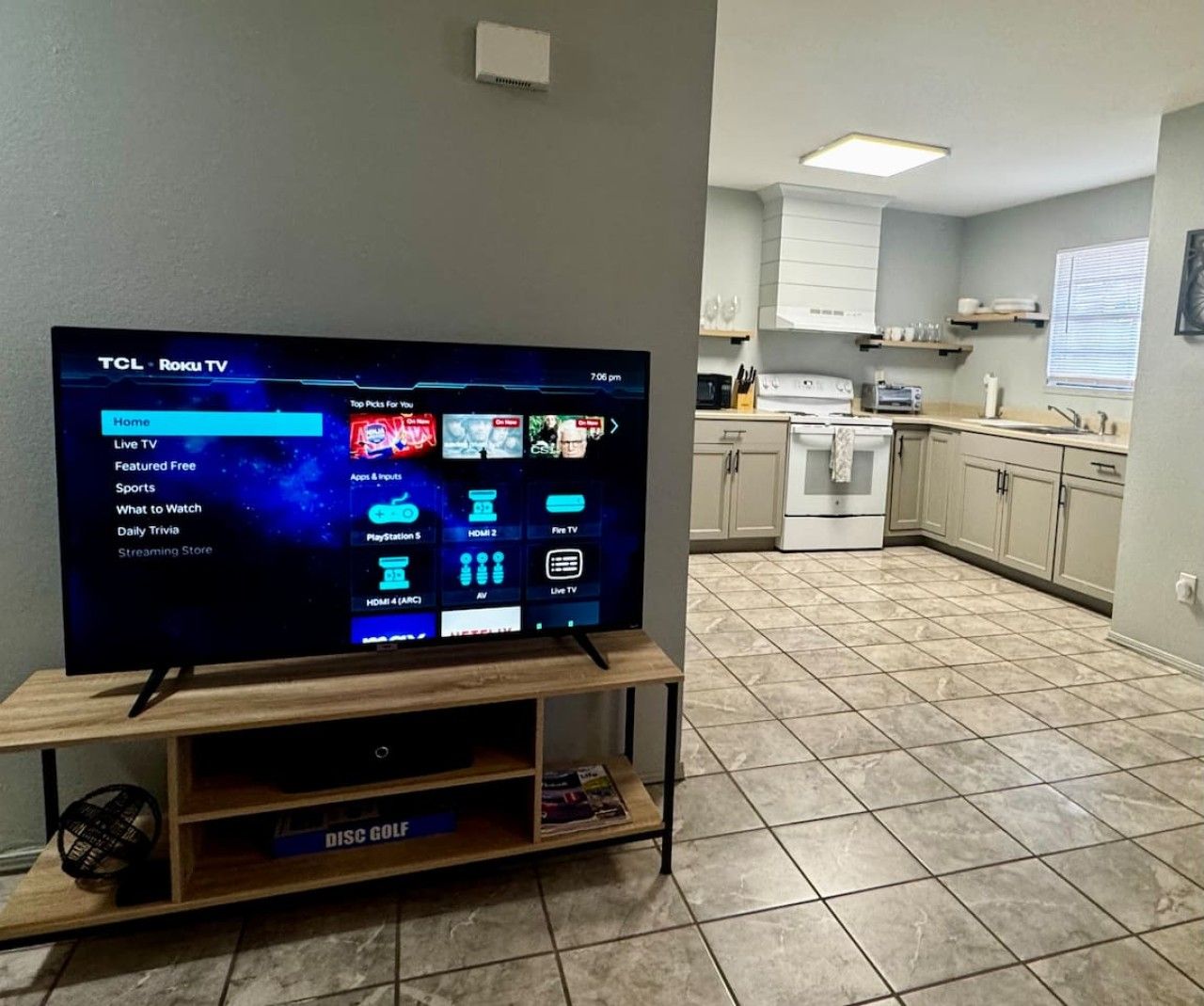 A TCL Roku TV on a wooden stand in a room with a tiled floor and a kitchen in the background.