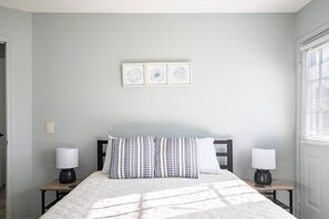 A bedroom with a bed, two bedside tables, a window with blinds, and three wall-mounted pictures.
