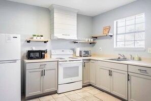A kitchen with a stove, microwave, and refrigerator. There are cabinets, a window with blinds, and a shelf with items.