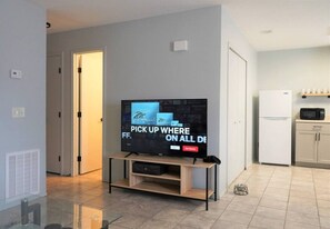 A modern living room with a flat-screen TV on a wooden stand, a kitchen area with a white refrigerator and microwave, and a tiled floor.