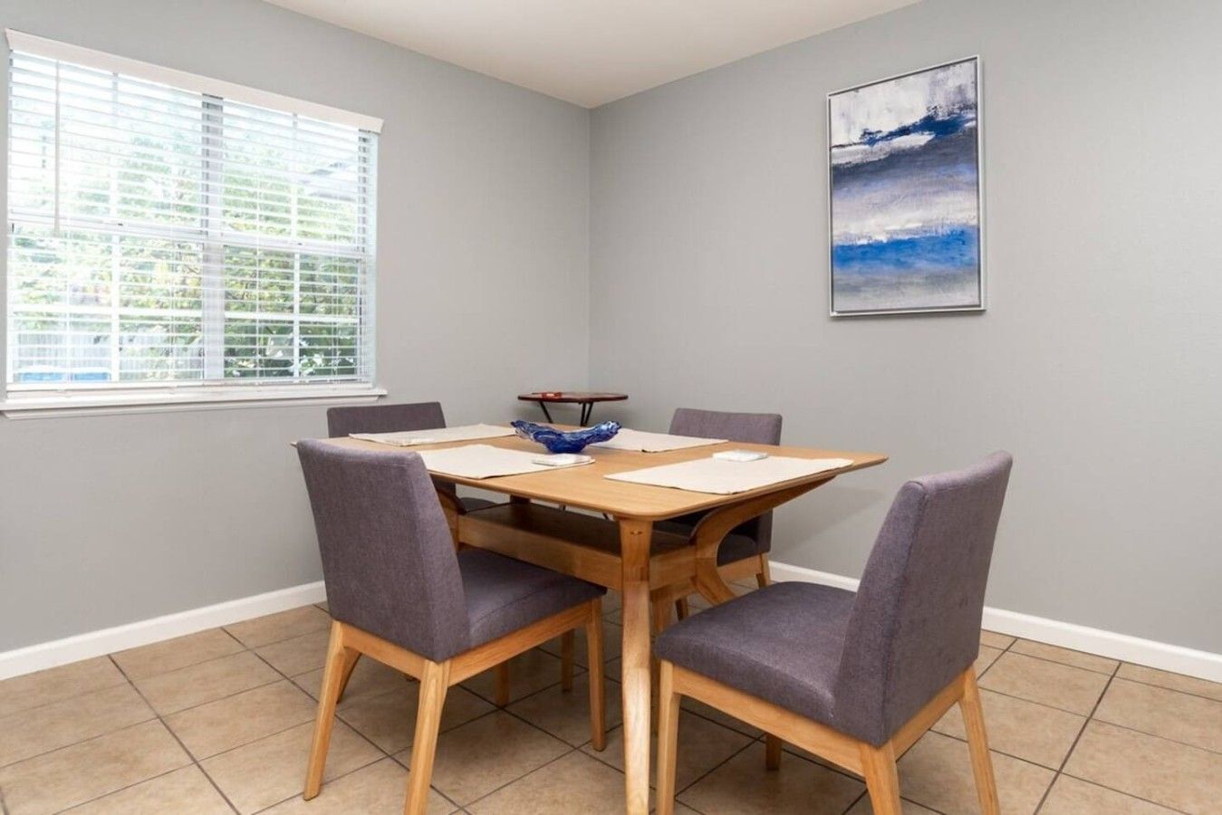 Un coin repas avec une table en bois, quatre chaises et une fenêtre équipée de stores.