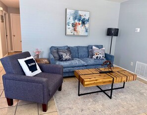 A living room with a sofa, armchair, coffee table, and artwork on the wall.