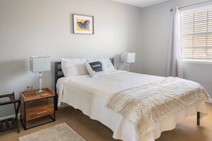 A bedroom with a bed, bedside lamps, a small wooden table, a chair, a rug, a framed picture on the wall, and a window with blinds.