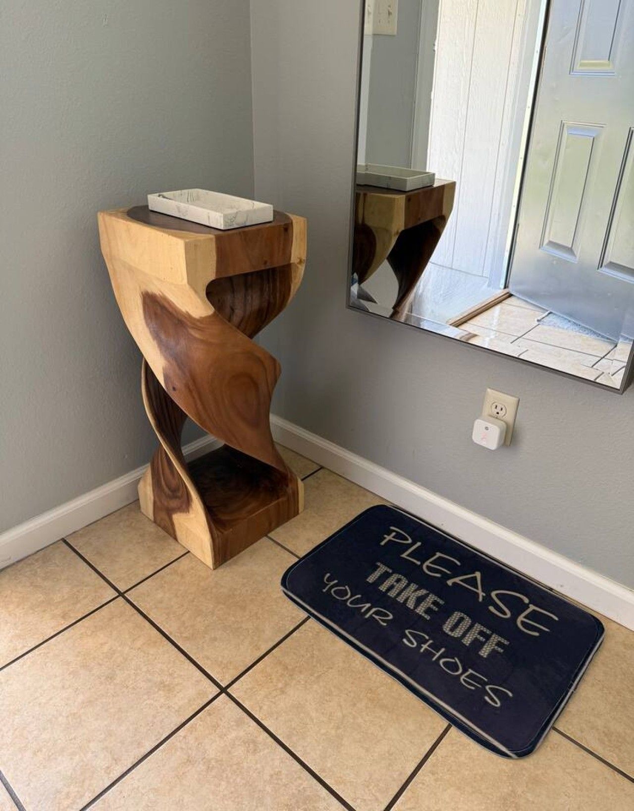 Et træ sidebord med en hvid genstand ovenpå, et spejl der afbilder en dør, og en måtte med teksten 'PLEASE TAKE OFF YOUR SHOES'.
