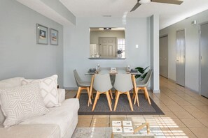 A modern living room with a sofa, dining table, and chairs. There is a kitchen area with cabinets and a refrigerator.
