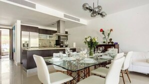 Ein moderner Essbereich mit Glastisch und weißen Stühlen, im Hintergrund eine Küche und ein weißes Sofa.