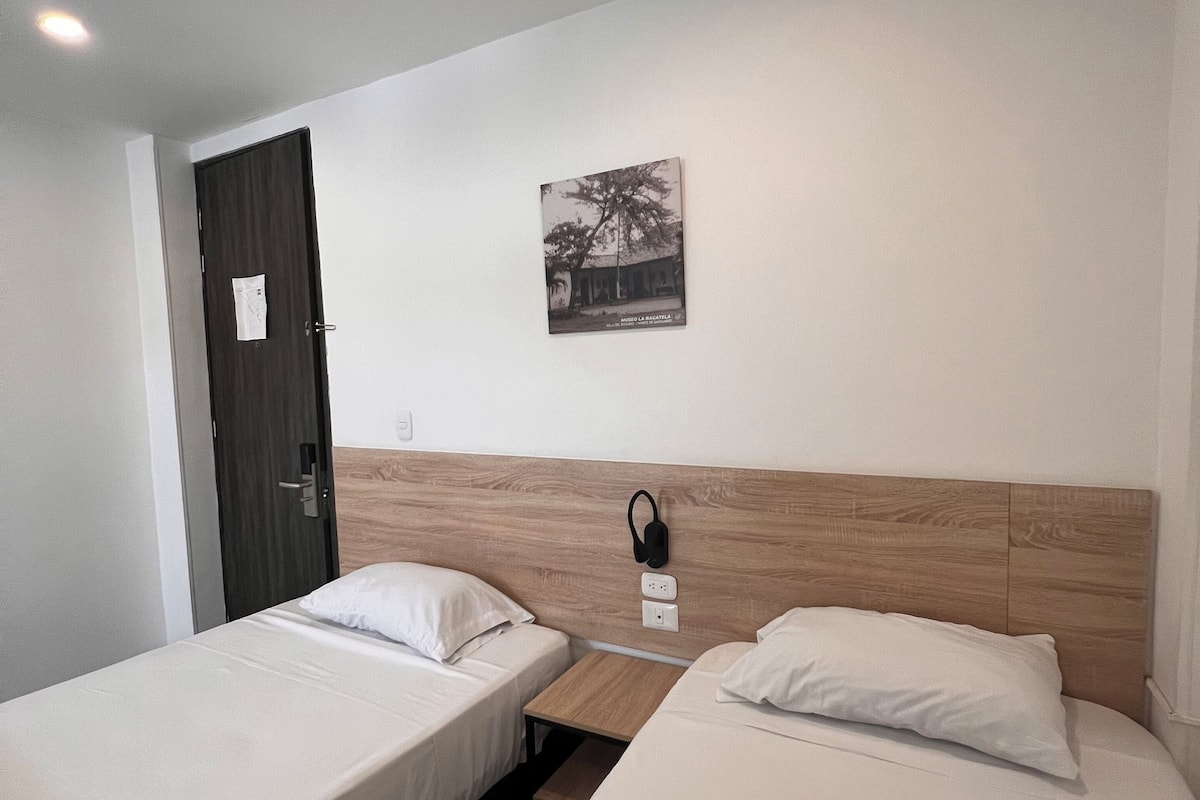 Habitación de hotel con dos camas individuales, cabecero de madera, una mesita de noche pequeña, un teléfono en la pared y una puerta con un papel adherido.