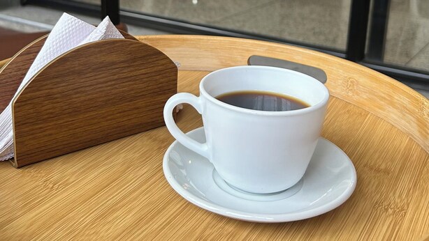 Una taza de café sobre una bandeja de madera con una servilleta, vista a través de una ventana que ofrece vistas de mesas al aire libre y árboles.