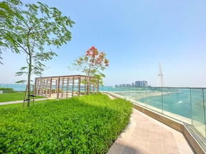 A landscaped area with a glass railing overlooking a body of water, a tree, and a modern structure. - Luxury StayCation - Incredible Sea View Apartment With Balcony (Dubai)