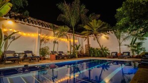 Una piscina con sillas de descanso e iluminación de guirnaldas, rodeada de palmeras y un muro blanco.