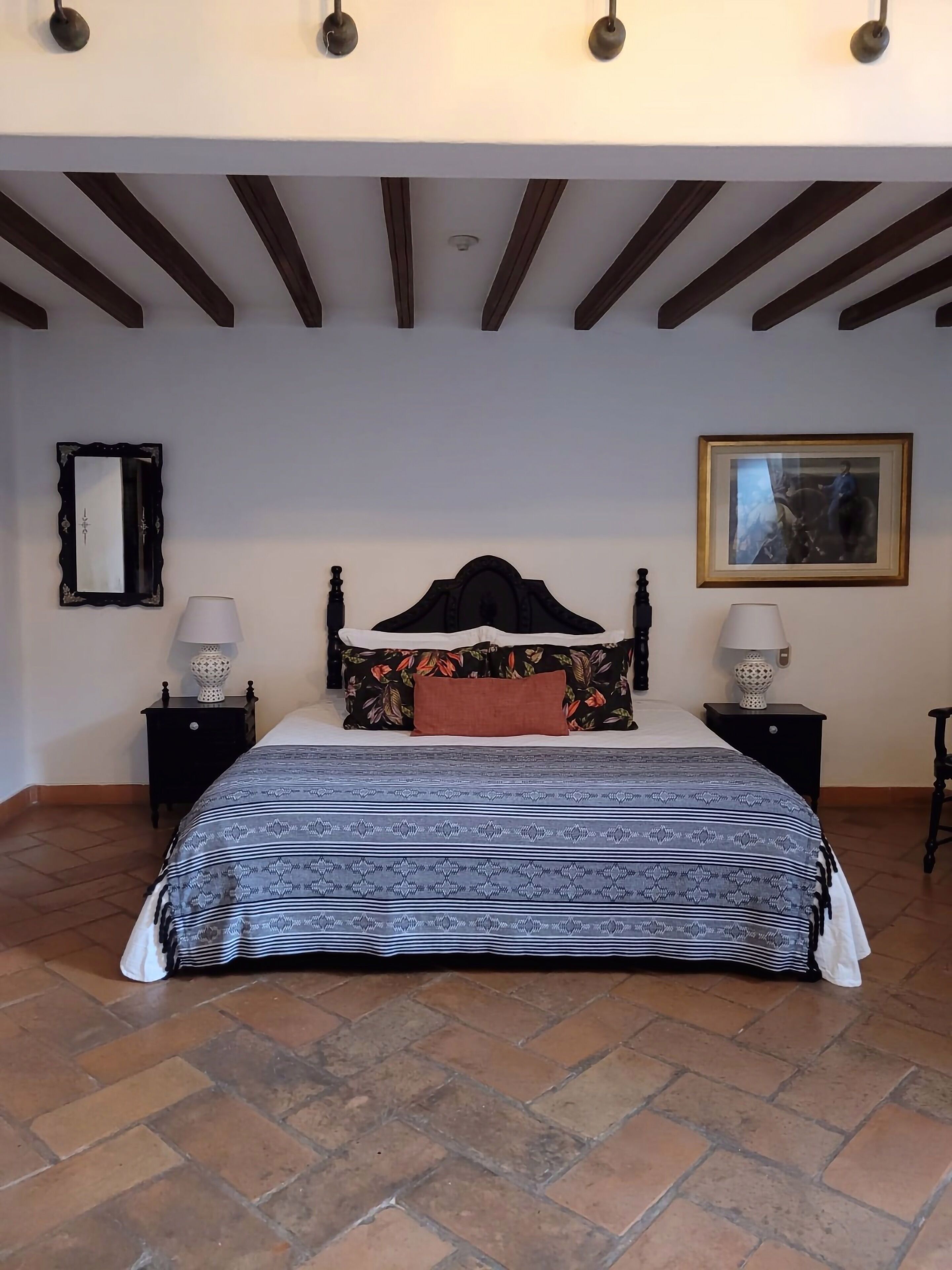 Un dormitorio con una colcha estampada, un cabecero de madera, dos mesitas de noche con lámparas, un espejo y un cuadro en la pared.