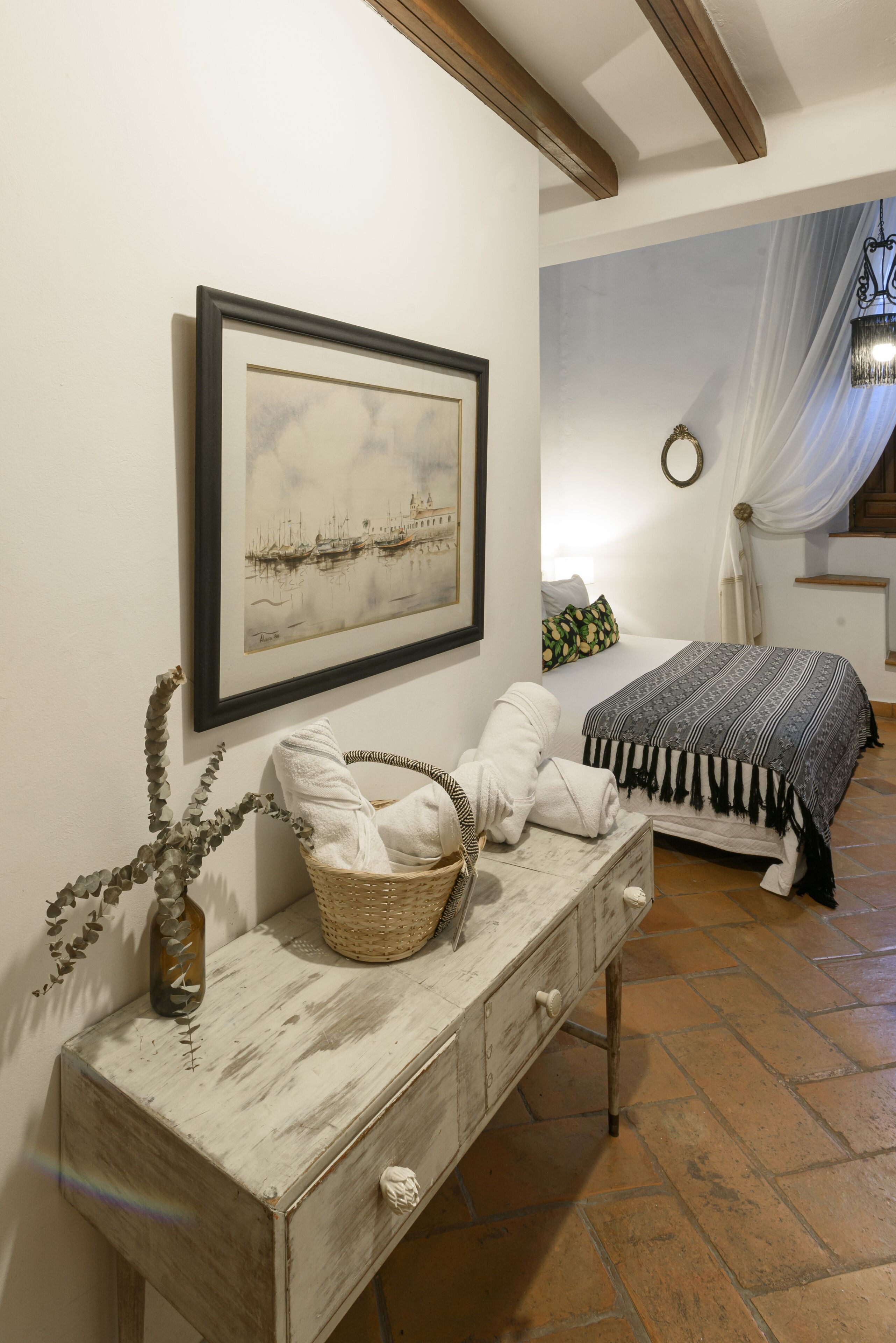 Un dormitorio con cama, una mesita de noche con cesta, un cuadro enmarcado y una escalera con cortina blanca.