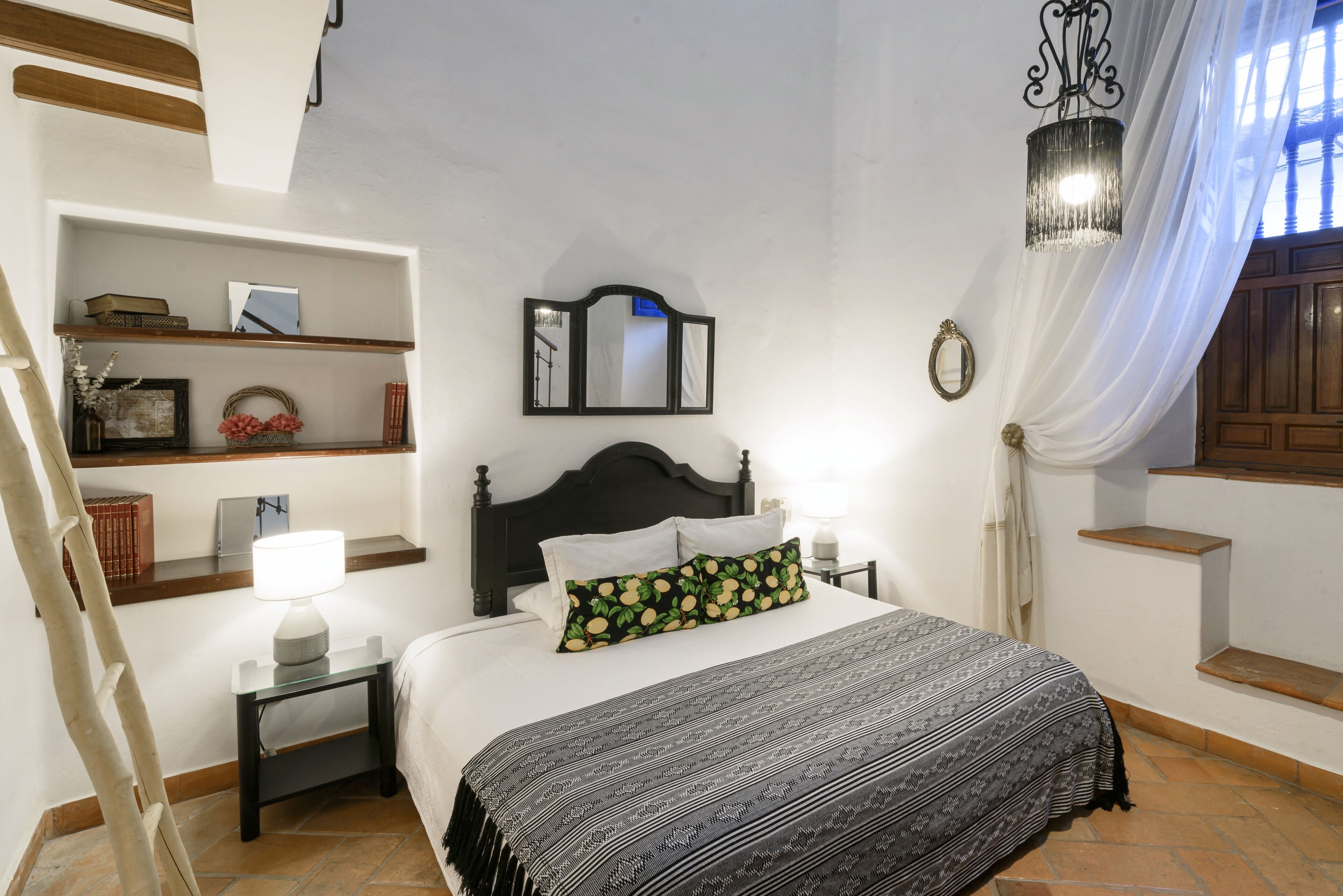 Un dormitorio con cama, mesita de noche, lámpara, espejo y estante con libros y objetos decorativos.