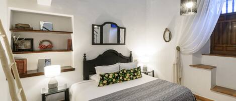 Un dormitorio con cama, mesita de noche, lámpara, espejo y estante con libros y objetos decorativos.