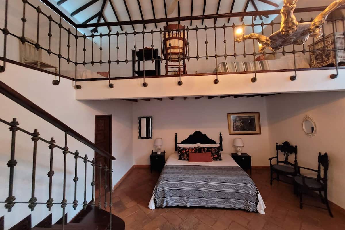 A bedroom with a large bed, a wooden floor, a balcony with a railing, and a ceiling fan.