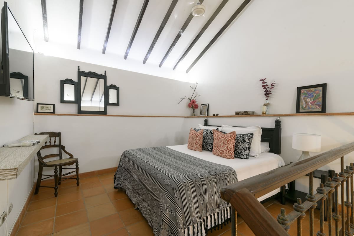 A bedroom with a bed, a desk, a chair, a mirror, and a window with a view of the sky.