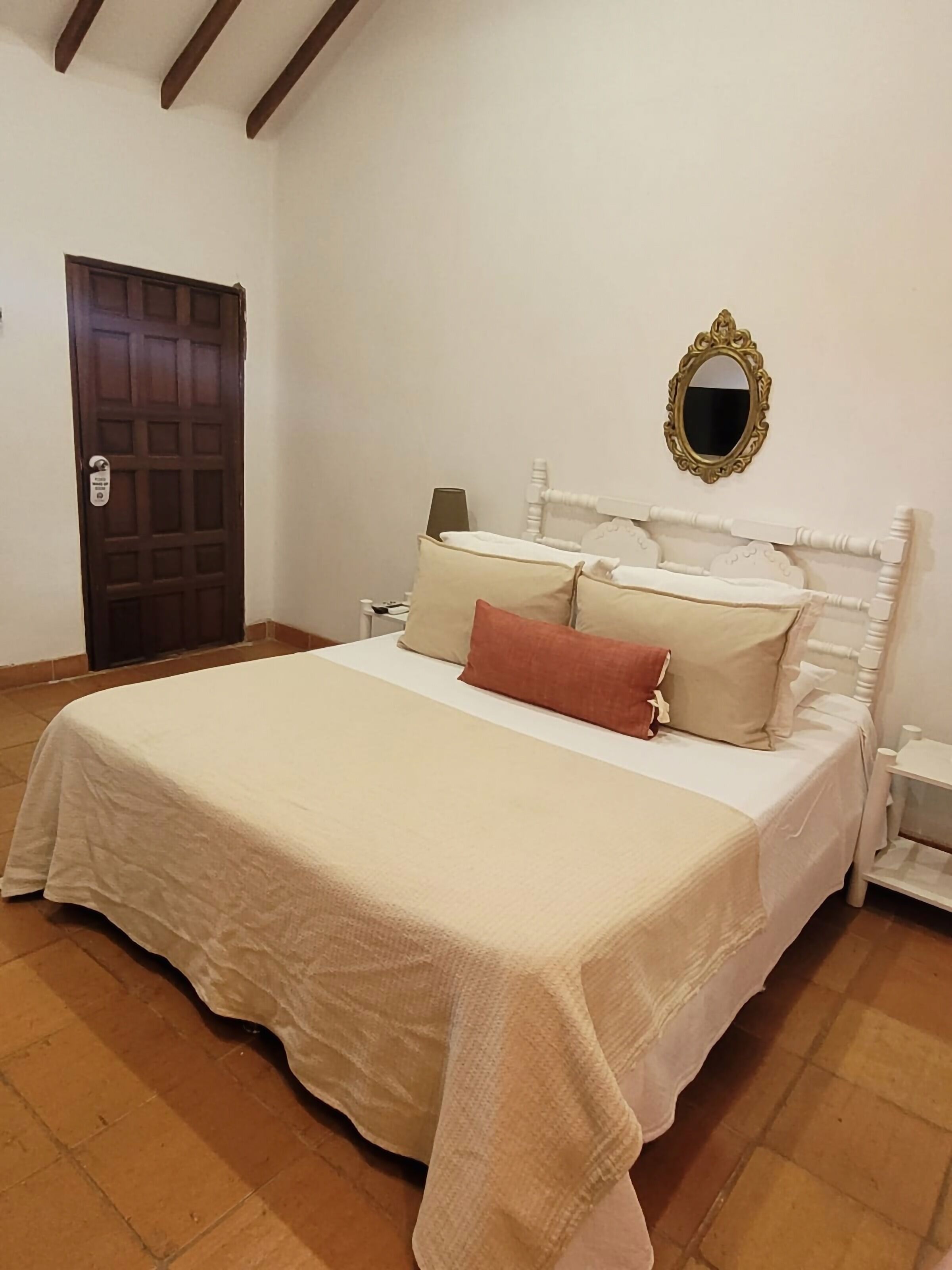 A neatly made bed with a white bedspread and pillows, a wooden door, a nightstand with a lamp, and a mirror on the wall.