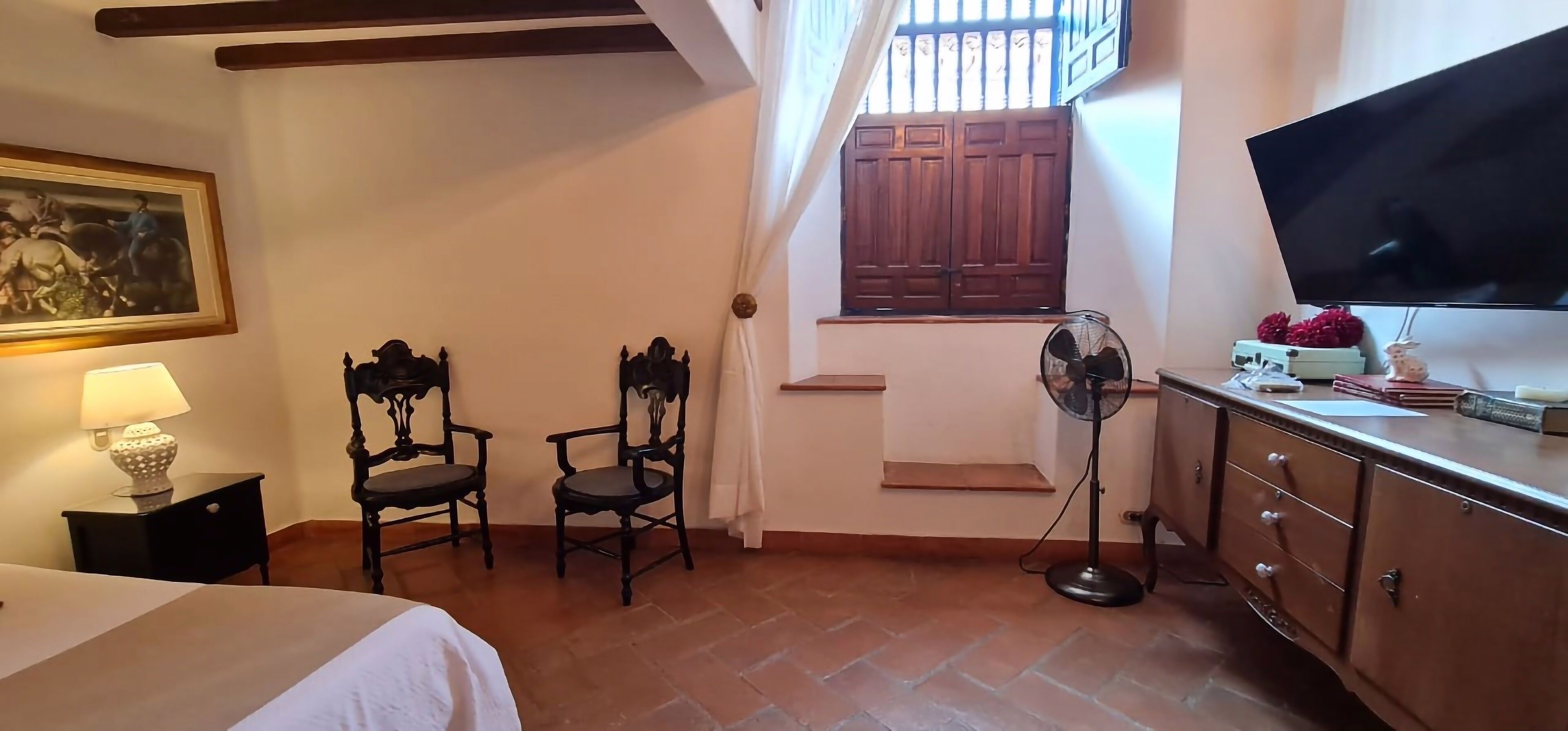 Habitación de hotel con cama, dos sillas, escritorio con televisor, ventilador y ventana con contraventanas de madera.