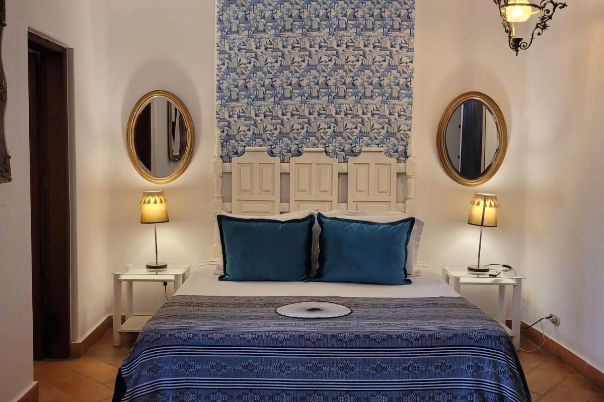 A bedroom with a patterned bedspread, two bedside tables with lamps, a headboard, and a tapestry on the wall.