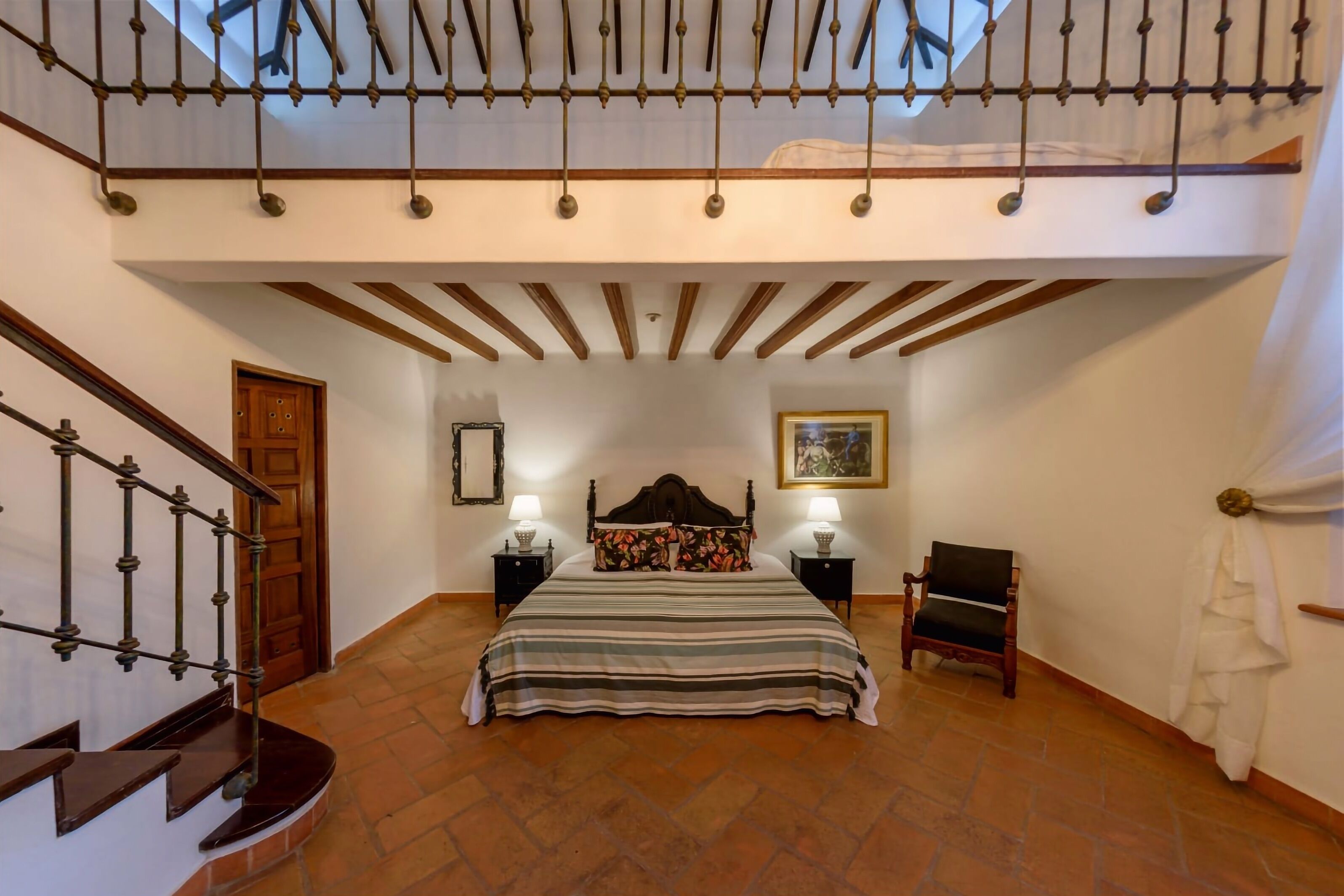 A bedroom with a bed, a chair, a nightstand, a picture on the wall, and a staircase.