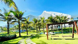 A well-maintained garden with a swing set, palm trees, and a paved pathway leading to a modern building with a unique architectural design. - Cam Ranh Nha Trang Seaview Condotel (Bac Cam Ranh)