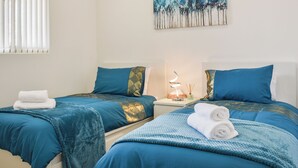 A hotel room with two beds, each with blue bedding and decorative pillows. There is a painting on the wall, a nightstand with a lamp, and a window with blinds. - A beautiful bungalow overlooking the coastal town of Llanelli. (Llanelli)