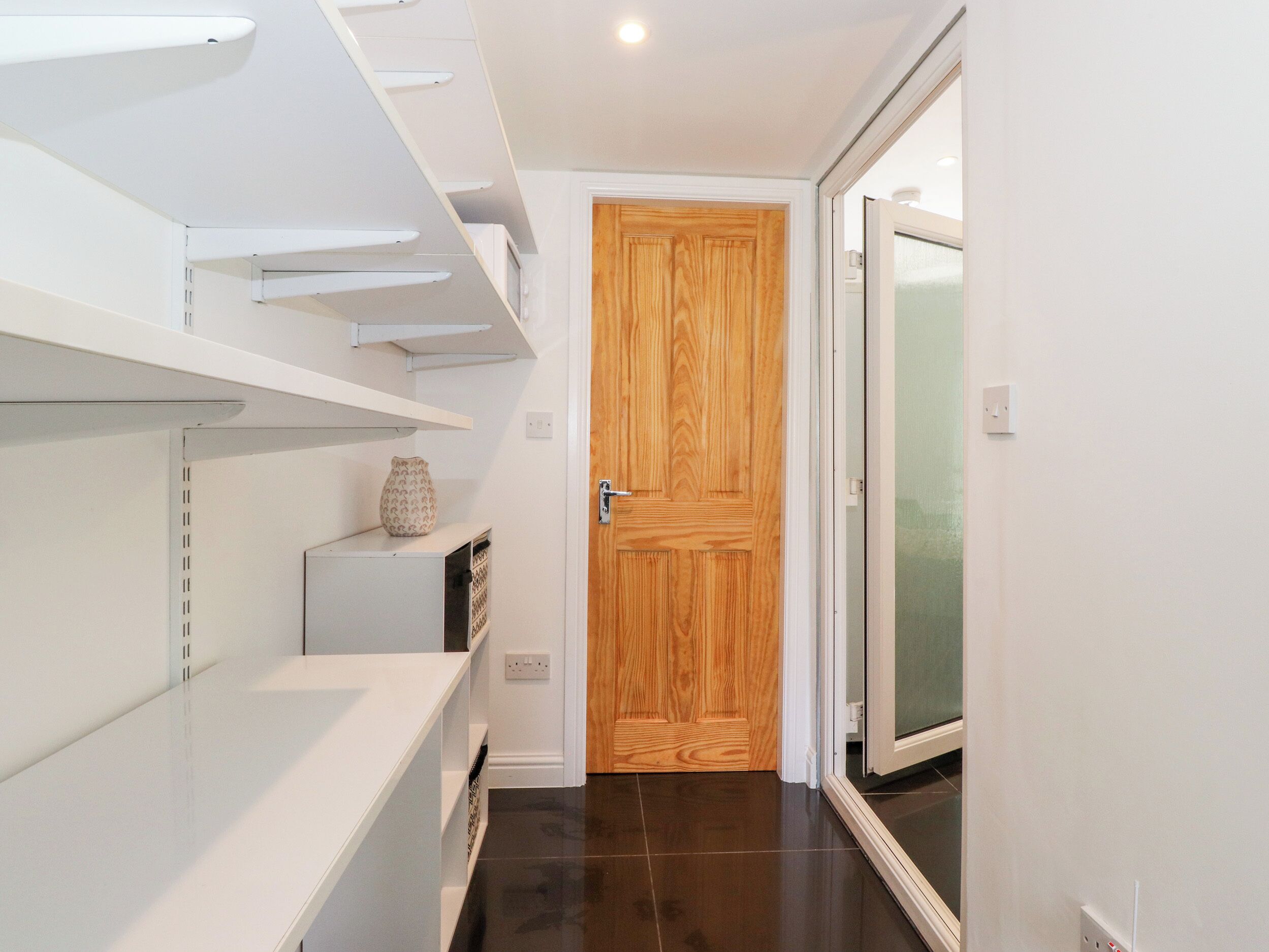 Un couloir avec des étagères blanches, une porte en bois et une porte en verre donnant sur une autre pièce.