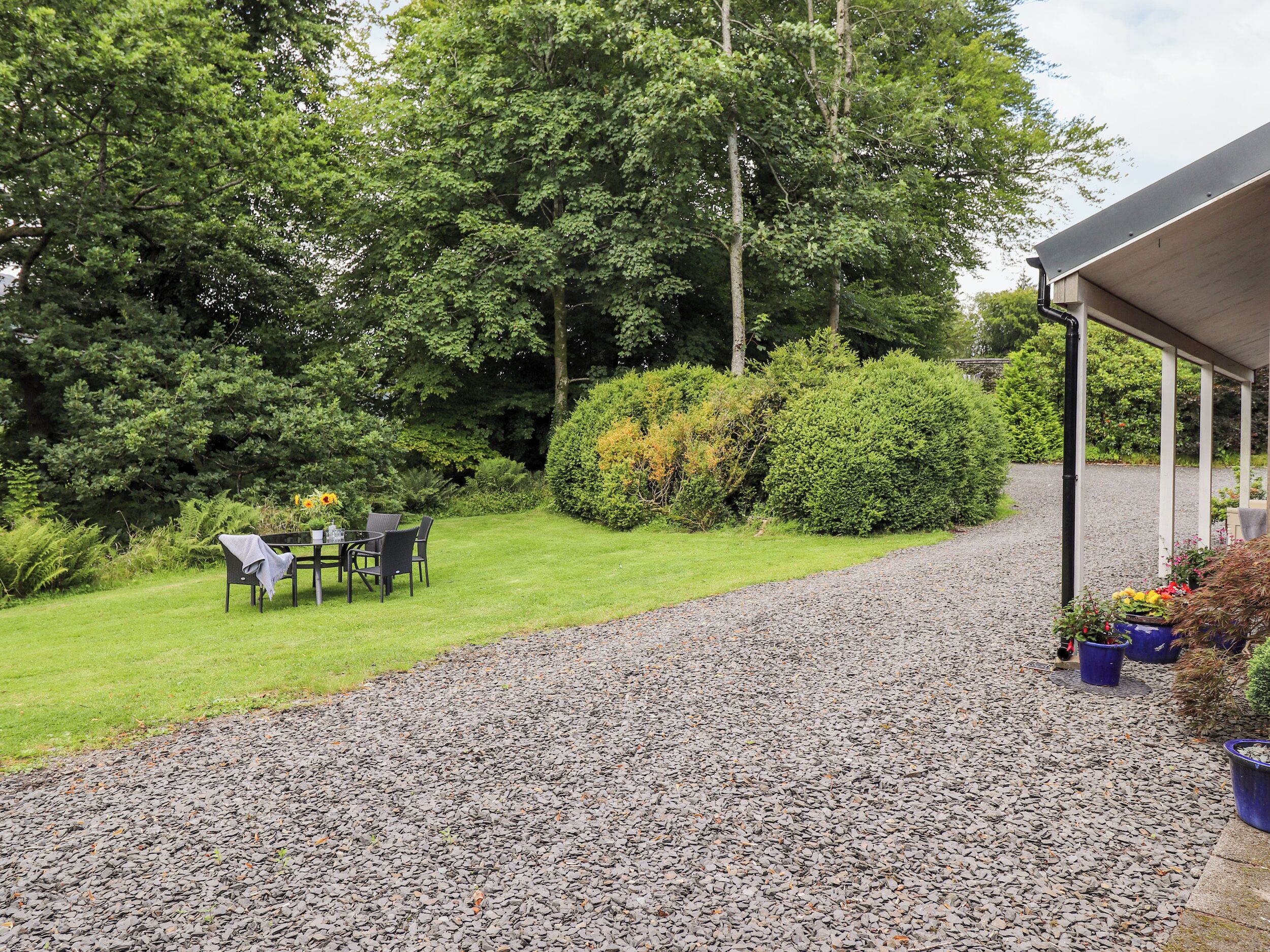 A gravel path leading to a covered outdoor seating area with a table and chairs, surrounded by lush greenery and trees.