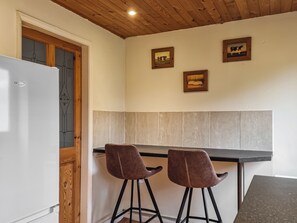 Ett kök med ett vitt kylskåp, två bruna barstolar, ett träpanelat tak och två inramade tavlor på väggen.