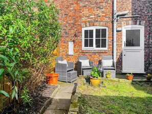 A brick building with a white door, a window, and a small outdoor seating area with wicker furniture and potted plants.