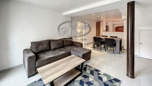 A modern living room with a sofa, a coffee table, and a dining area with chairs and a kitchen in the background.