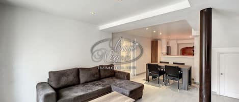 A modern living room with a sofa, a coffee table, and a dining area with chairs and a kitchen in the background.