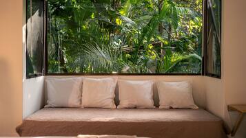 A bed with a beige mattress and white pillows, positioned in front of a window with a view of dense green foliage.