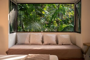 A bed with a beige mattress and white pillows, positioned in front of a window with a view of dense green foliage. - Pájaros · Bright Jungle Cabin + Pool · Walk to Beach · Sleeps 3 (Cóbano)