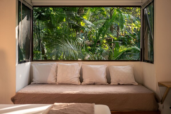 A bed with a beige mattress and white pillows, positioned in front of a window with a view of dense green foliage.