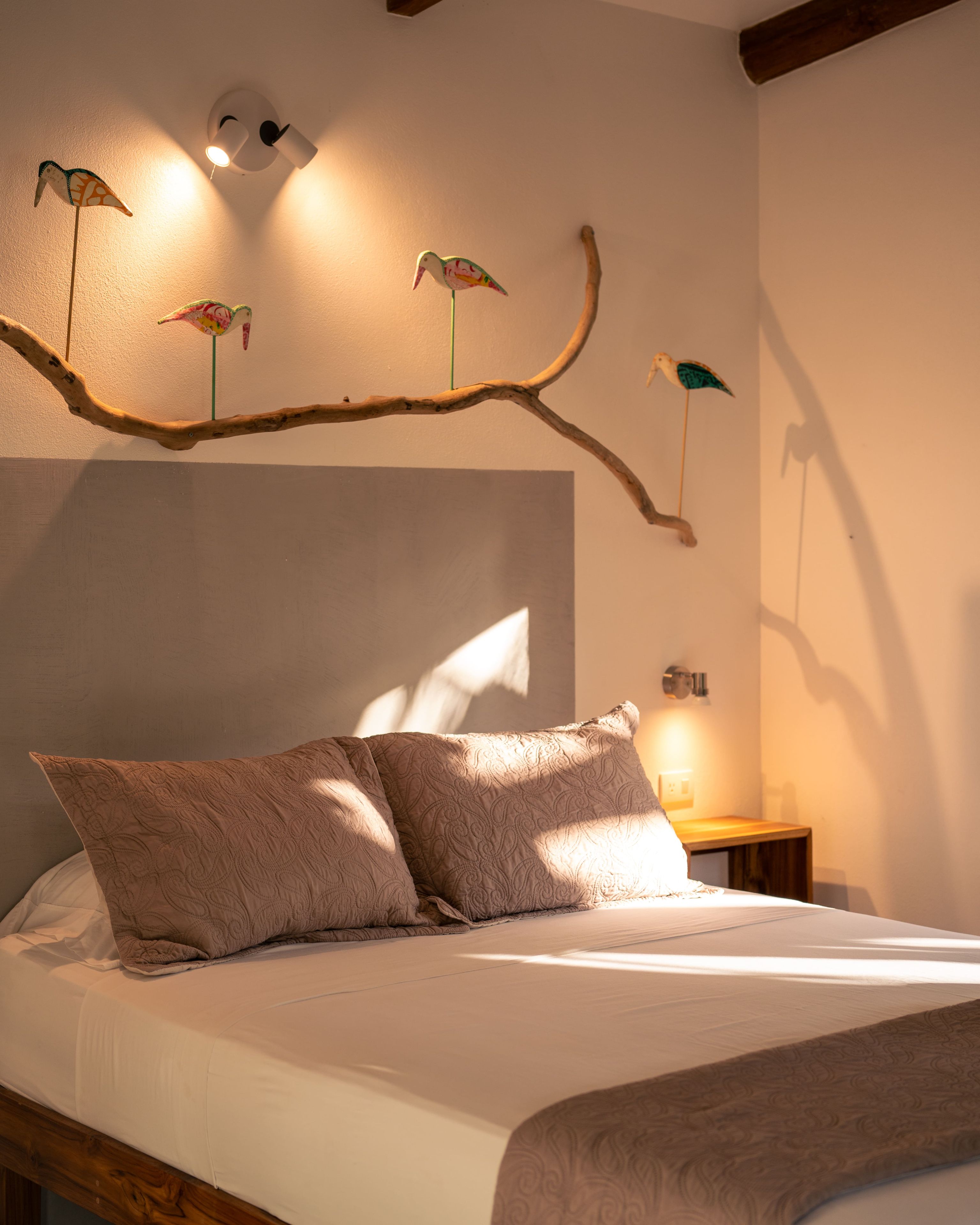 A bedroom with a bed, pillows, a branch with hanging bird decorations, and a wall-mounted light fixture.