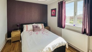 A bedroom with a bed, a nightstand, a window with curtains, and a radiator. - 2 bedroom accommodation in Gramsdale, Isle of Benbecula (Gramsdale, Isle of Benbecula)