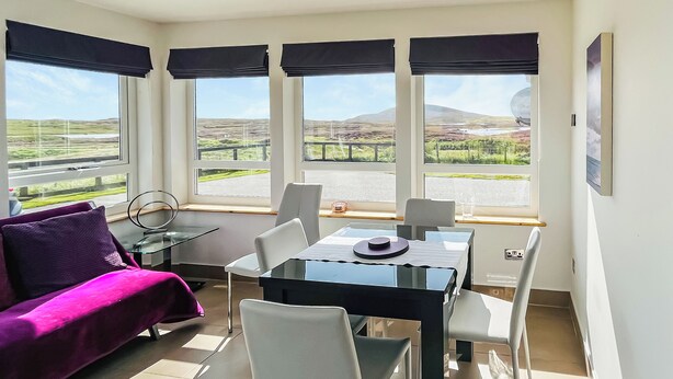 A modern room with a dining table, chairs, a purple sofa, and large windows offering a view of the countryside. - 2 bedroom accommodation in Gramsdale, Isle of Benbecula (Gramsdale, Isle of Benbecula)