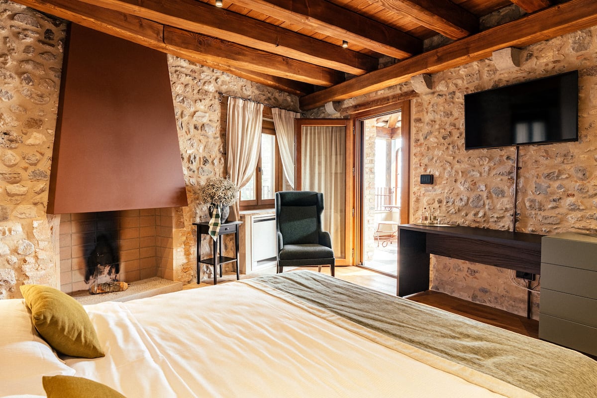 Ein Schlafzimmer mit Steinstruktur, Holzbalken, Kamin, Bett mit Kissen, Sessel und Fernseher.
