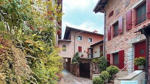 A narrow alleyway with stone buildings, red shutters, and lush greenery. - Casa del Vicario (Fagagna)