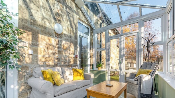 A conservatory with a stone wall, a sofa with yellow cushions, a wooden table, and a wicker chair. - 2 bedroom accommodation in Hexham (Hexham)