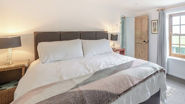 Quarto com uma cama grande, uma mesa de cabeceira com abajur, uma porta de madeira e uma janela com cortinas.