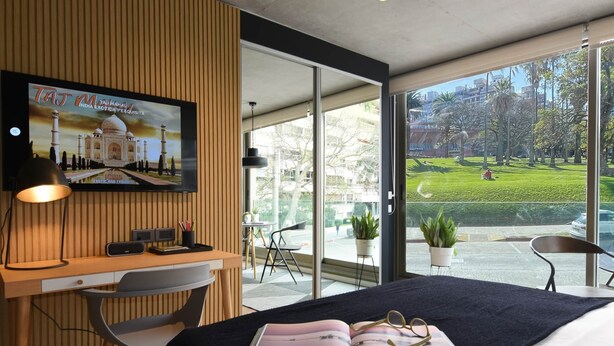 Een moderne hotelkamer met een groot bed, een bureau met een computer, een televisie waarop de Taj Mahal te zien is en uitzicht op een park en gebouwen.