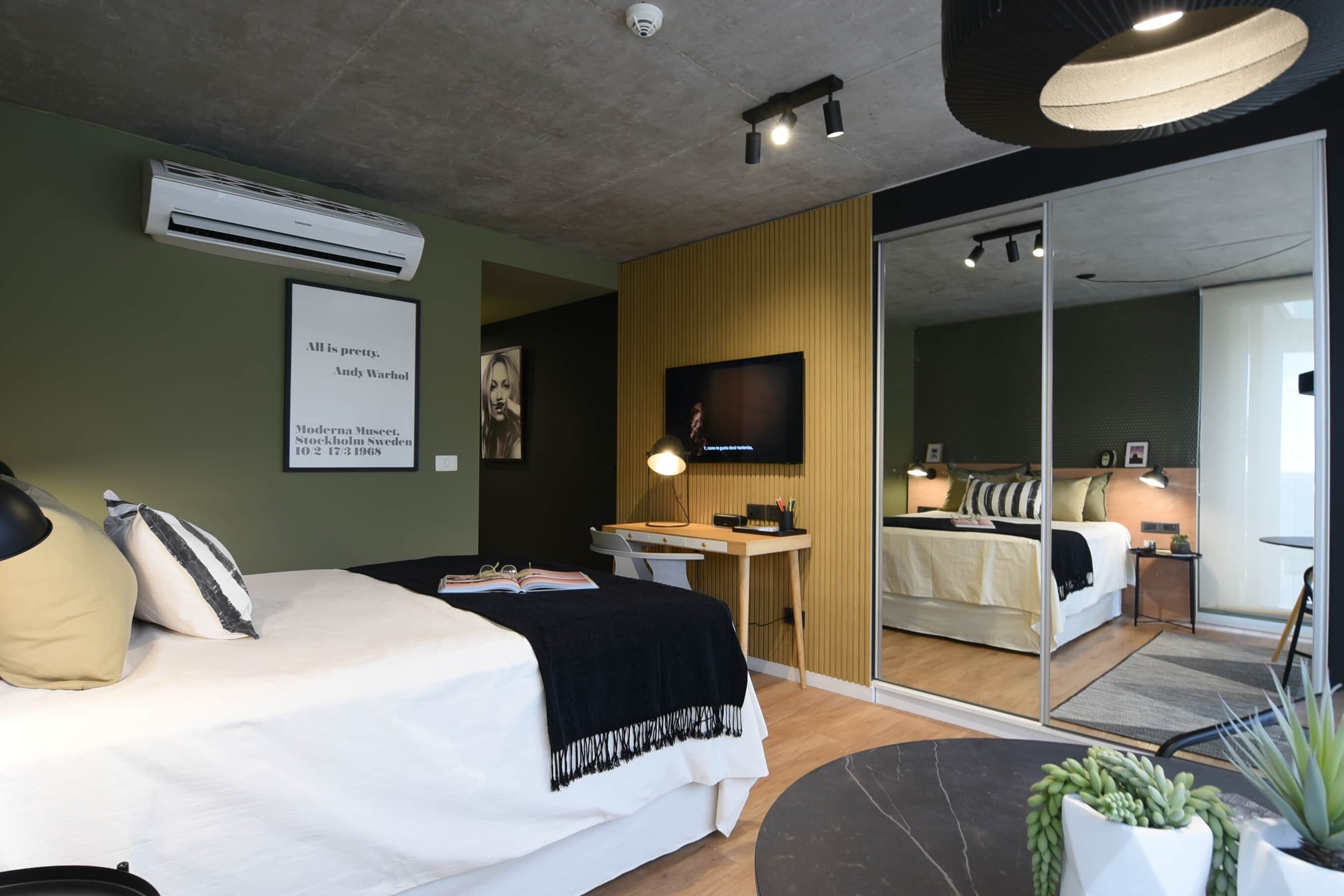 A modern hotel room with a bed, desk, and a large mirror reflecting another room.